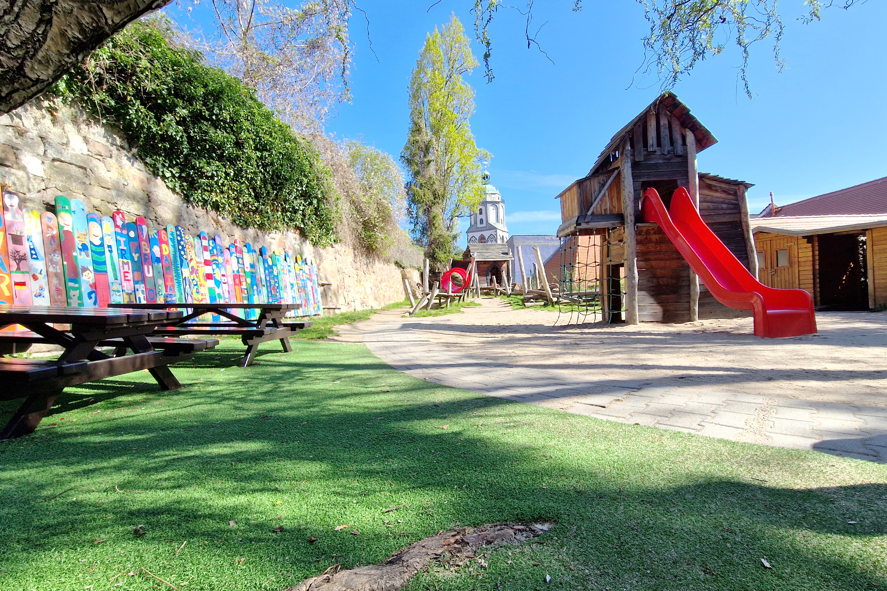 Spielbereich mit Kunstrasen, Sandsteinmauer, Rutsche, bunten Zaunslatten