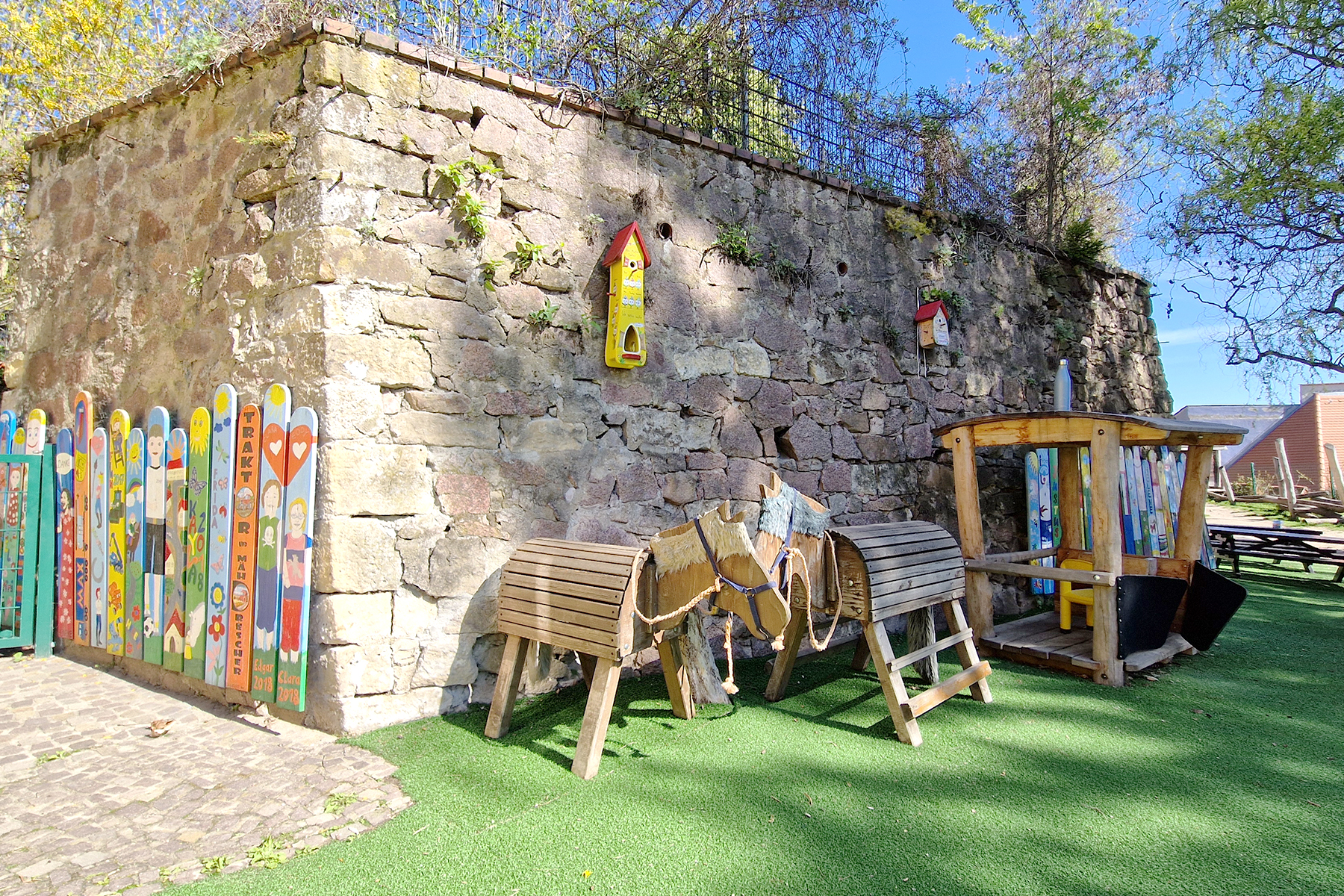 Außenspielbereich mit Kunstrasen, Sandsteinmauer, bunt bemalten Zaunslatten, Spielpferden